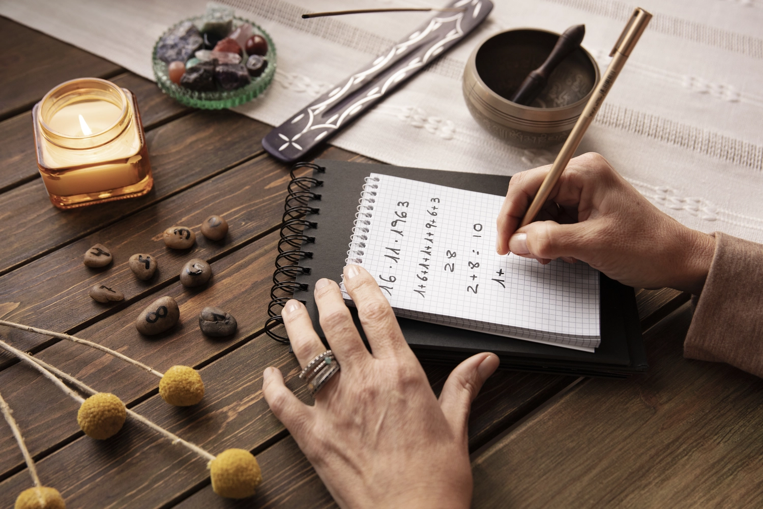 NumeroWunder - Numerologin und Thetahealing Praktikerin - Eine Person notiert numerologische Berechnungen in einem Notizbuch auf einem rustikalen Holztisch, umgeben von einer Kerze, Kristallen und einer Klangschale.