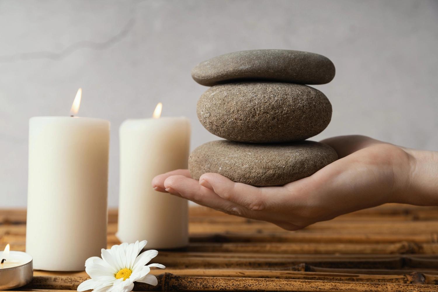 NumeroWunder - Numerologin und Thetahealing Praktikerin - Eine Hand balanciert drei glatte, graue Spa-Steine neben brennenden weißen Stumpenkerzen und einer weißen Gänseblümchenblüte.